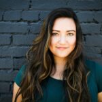 A captivating portrait of a woman smiling confidently in front of a brick wall, showcasing her long brunette hair and vibrant eyes.